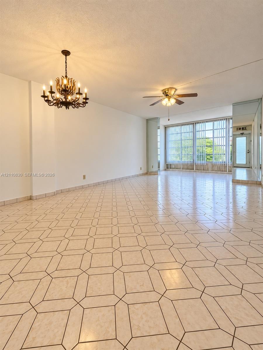 Spacious interior featuring patterned tile flooring, a multi-arm candelabra-style chandelier, a ceiling fan with integrated lighting, and floor-to-ceiling windows with vertical blinds Spacious interior featuring patterned tile flooring, a multi-arm candelabra-style chandelier, a ceiling fan with integrated lighting, and floor-to-ceiling windows with vertical blinds