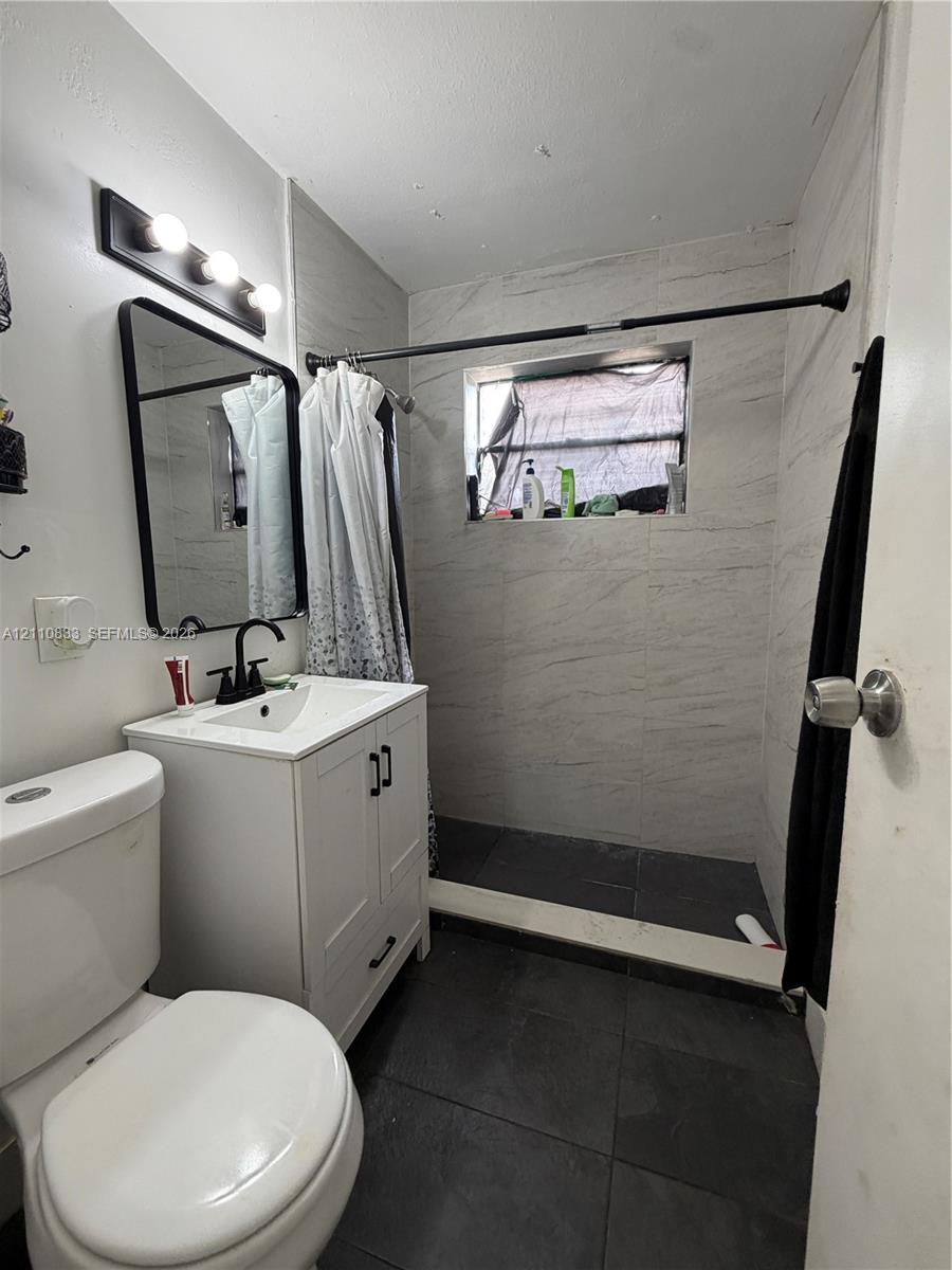 Bathroom featuring a white vanity cabinet with a stone-top integrated sink, matte black faucet, wall-mounted mirror, three-light vanity fixture, and tiled shower surround
