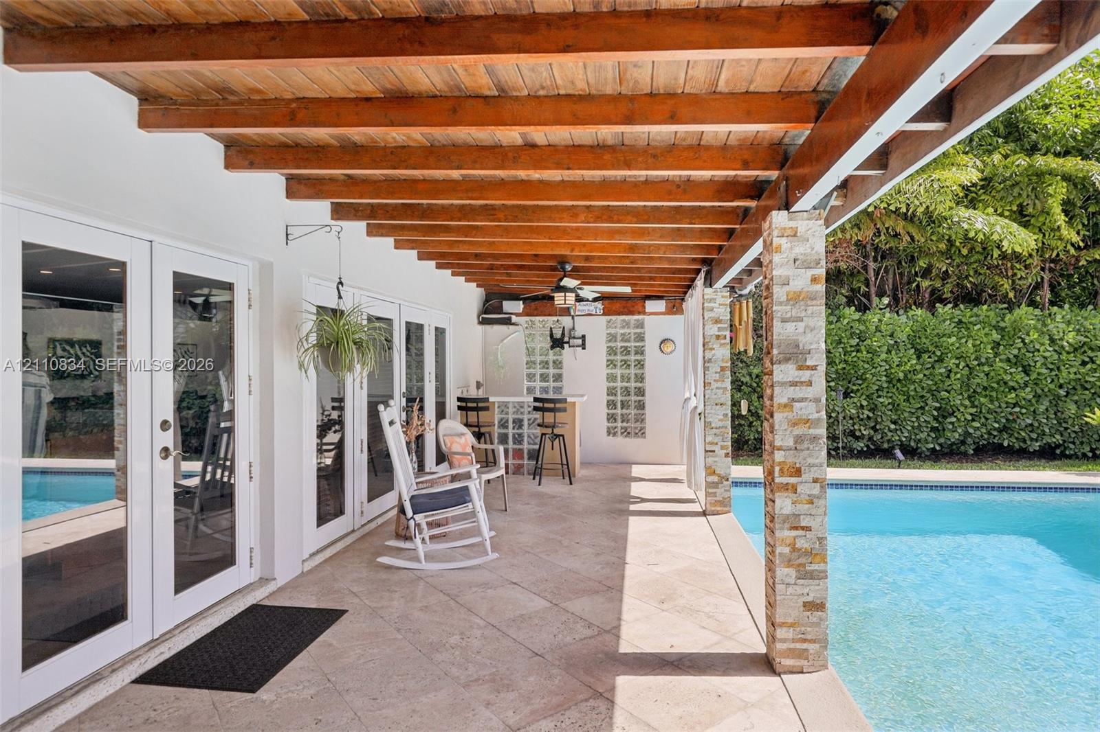 Covered patio featuring a wood-beam pergola, stone-clad support columns, natural stone flooring, and built-in glass block bar, complemented by a swimming pool
