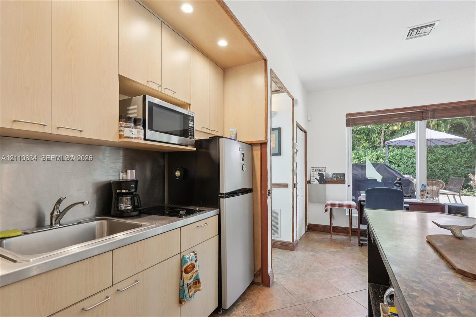 Kitchenette featuring light wood-finish cabinetry, stainless steel sink with arched faucet, metallic backsplash, built-in microwave, and tile flooring