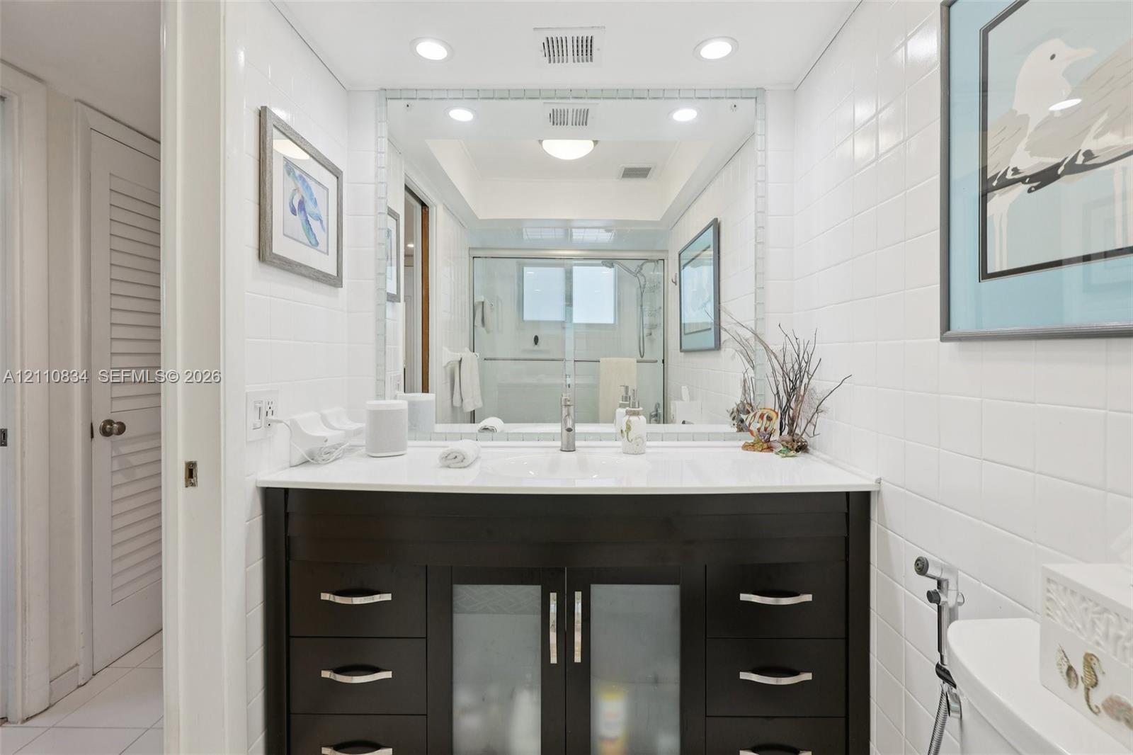 Bathroom featuring a dark wood-finish vanity with an integrated basin, white solid-surface countertop, recessed lighting, and a floor-to-ceiling mirror with a tiled border