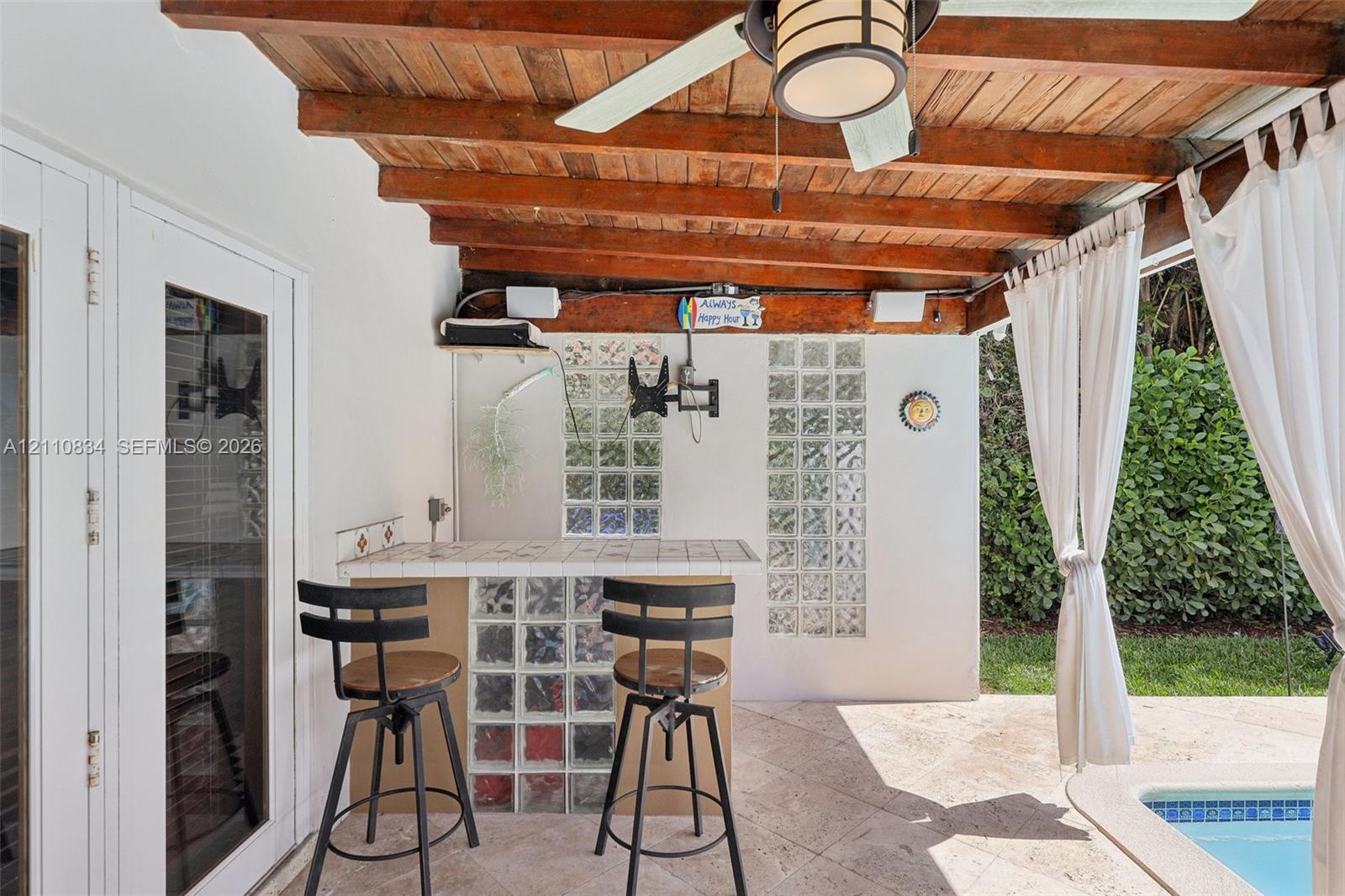 Covered patio featuring an exposed wood-beam ceiling, ceiling fan with integrated lighting, tiled bar with glass-block base, and stone-finish flooring