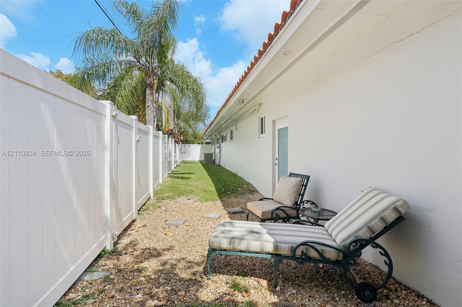 Side exterior featuring stucco siding, clay tile roof eaves, white vinyl privacy fencing, and a gravel ground cover