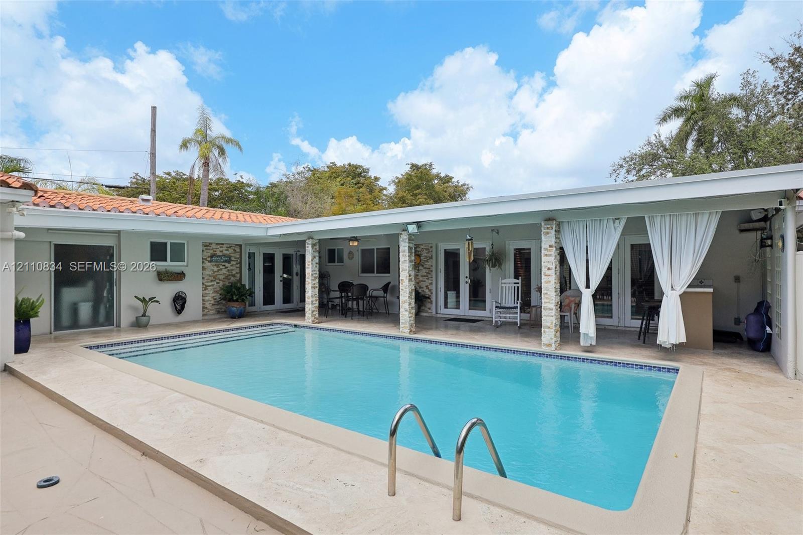 In-ground swimming pool with stone-tiled decking and covered patio area featuring stone-clad support columns and light-toned exterior finish