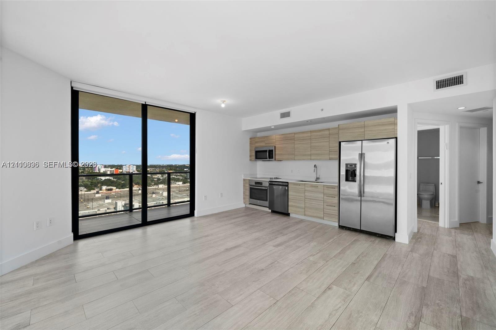 Open-concept living space featuring wood-finish flooring, floor-to-ceiling glass sliding doors, horizontal wood-grain cabinetry, stainless steel appliances, and a private balcony