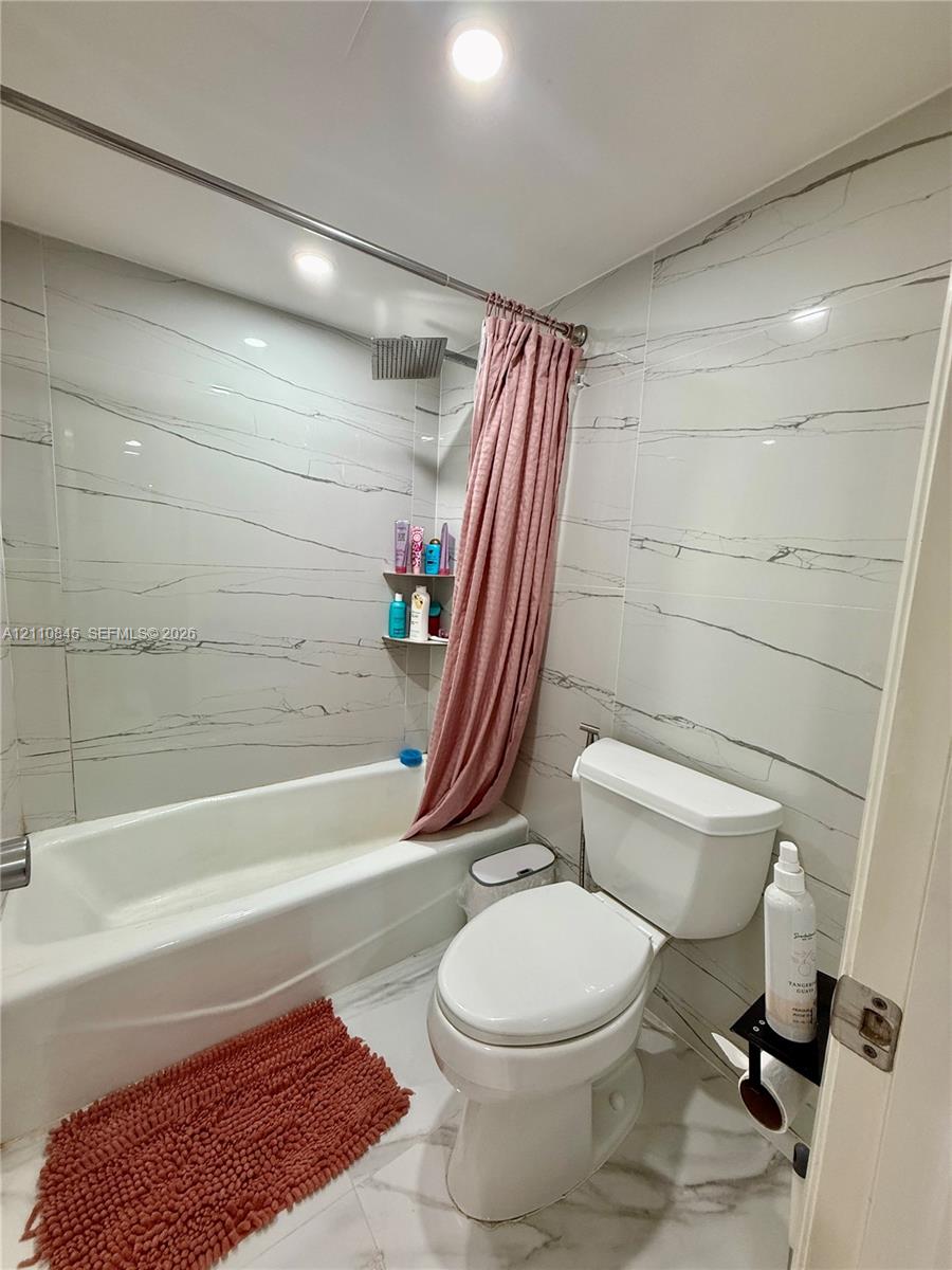 Full-height wall tiling featuring veined detailing, white acrylic bathtub, integrated shower head, recessed ceiling lighting, and large-format tile flooring