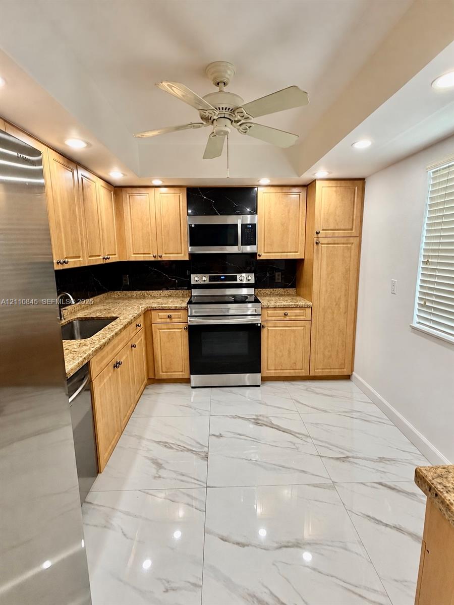 Kitchen featuring light wood cabinetry, stone countertops, stainless steel appliances, a dark stone backsplash, and large-format tile flooring