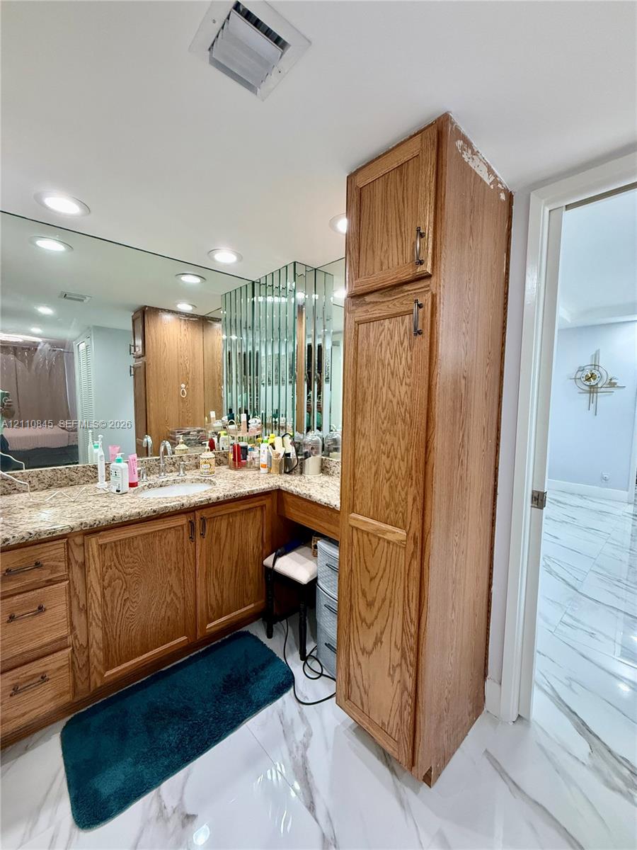 Vanity featuring wood cabinetry, natural stone countertop, recessed lighting, integrated full-wall mirror, and floor-to-ceiling storage cabinet