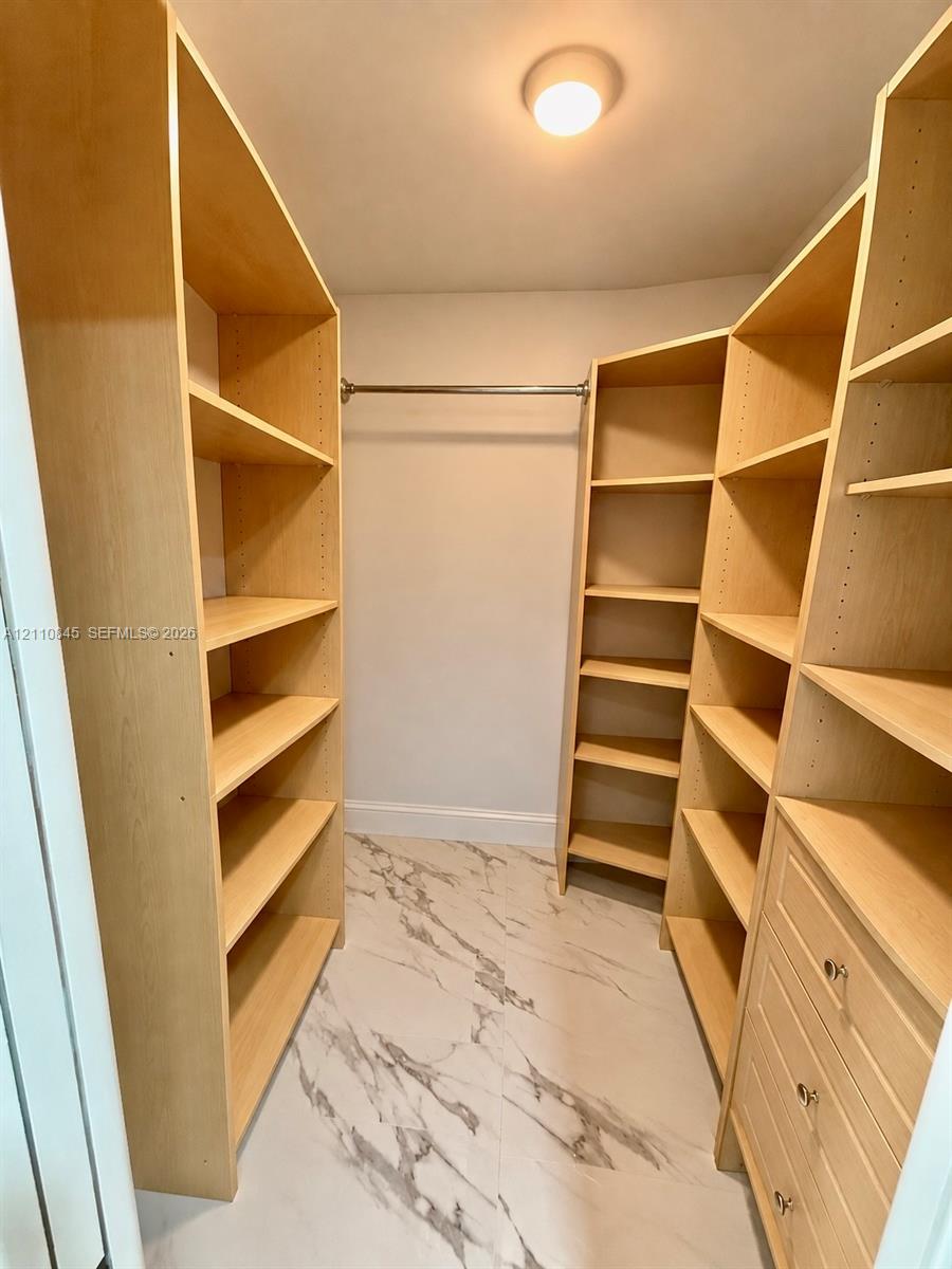 Walk-in closet featuring light-toned custom shelving, integrated drawer stack, metal hanging rod, and large-format tile flooring with a marble-patterned finish