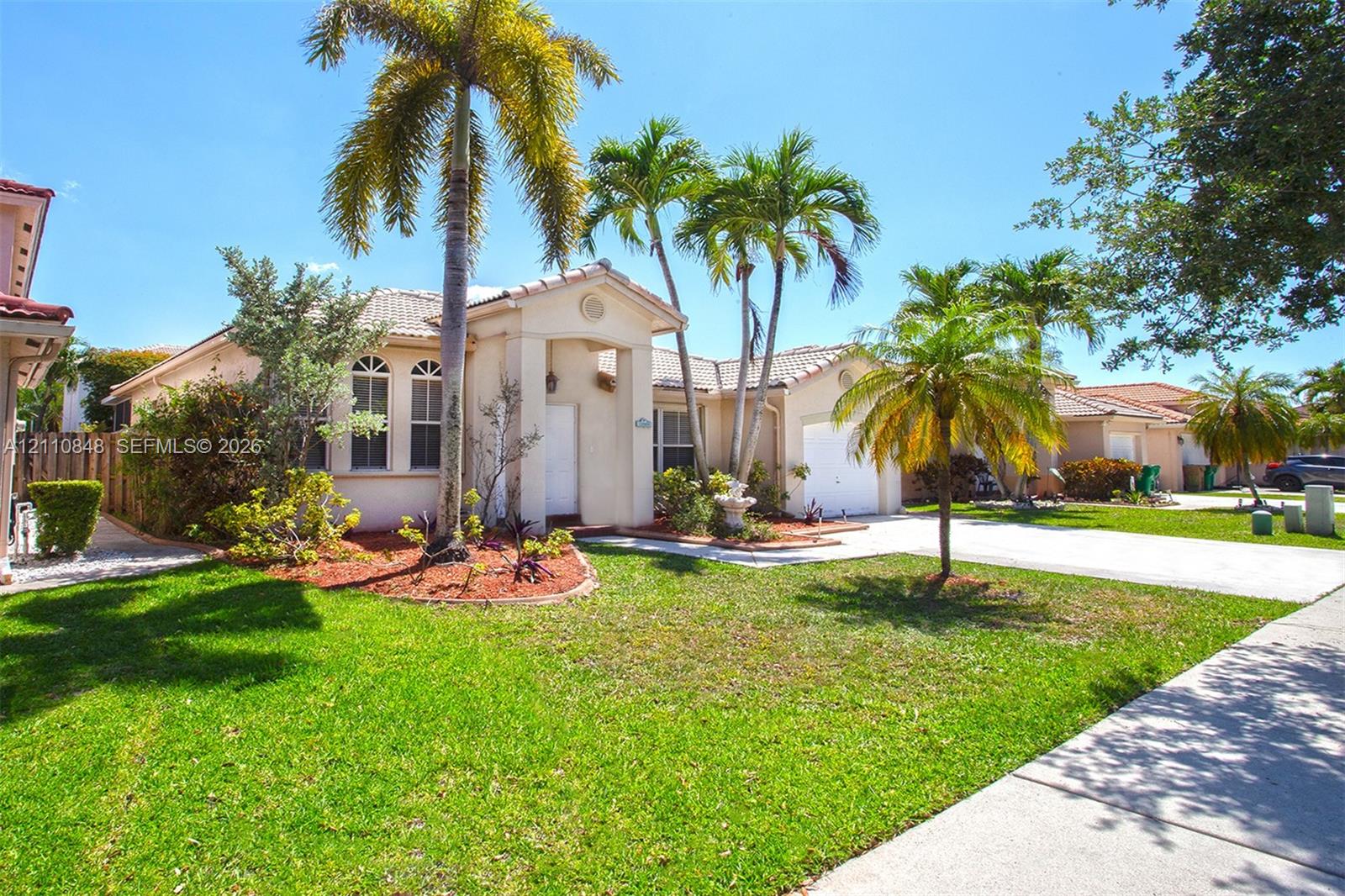 Stucco exterior residence featuring a tile roof, arched windows, covered entryway, and an attached two-car garage set on a landscaped lot with mature palm trees