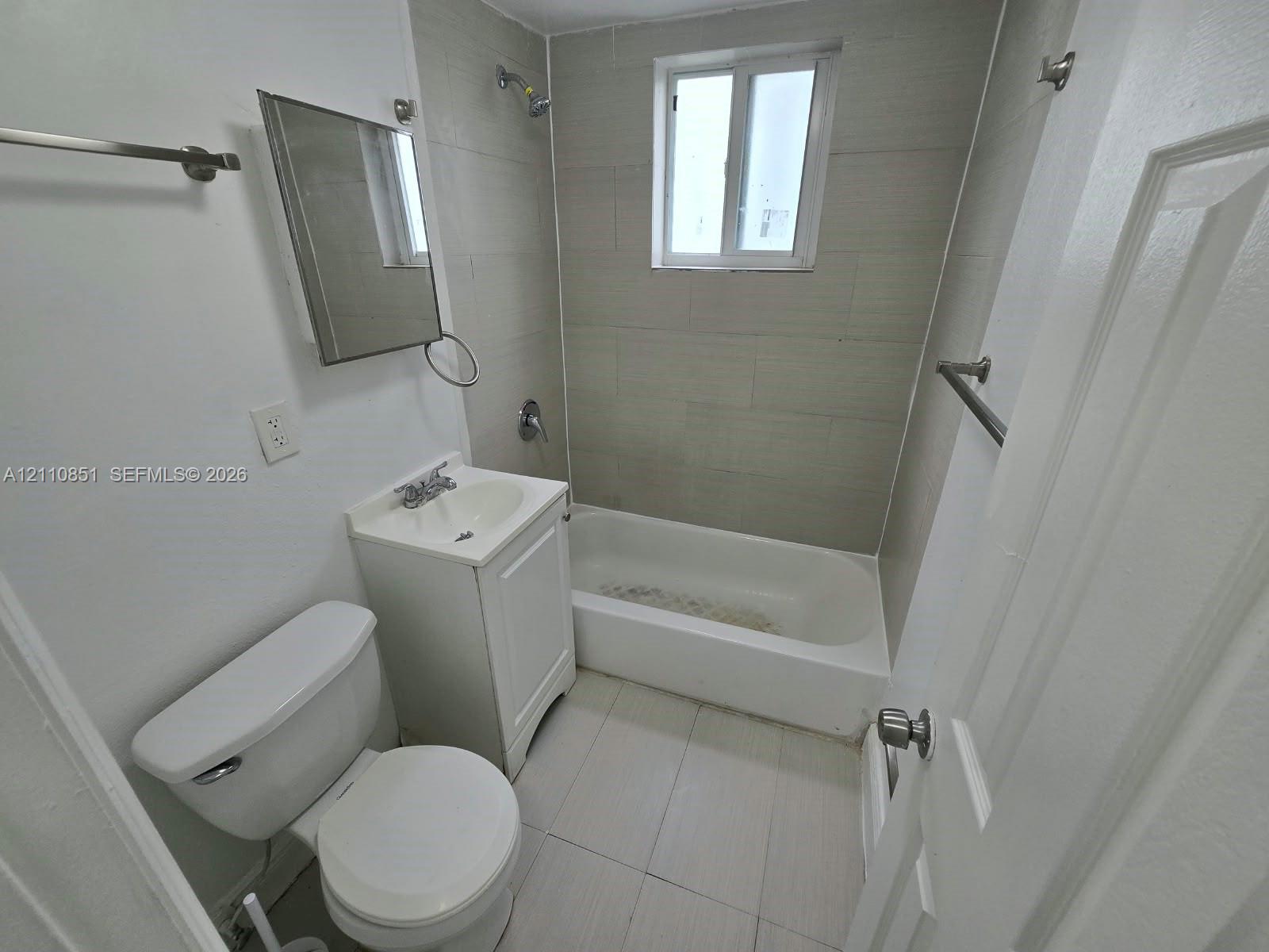 Bathroom featuring a white pedestal vanity, ceramic tile flooring, a tub-shower combination with tile surround, a rectangular wall-mounted mirror, and a single-pane window