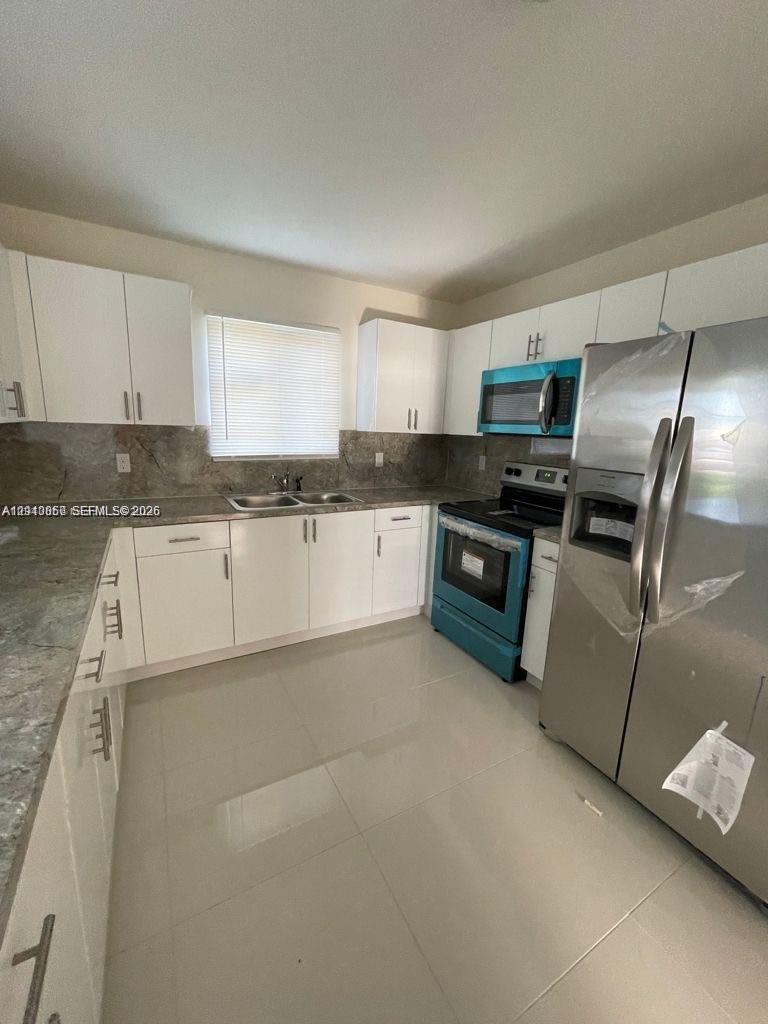 Kitchen featuring white shaker-style cabinetry, stone-surface countertops, stone-tile backsplash, stainless steel appliances, and large-format tile flooring