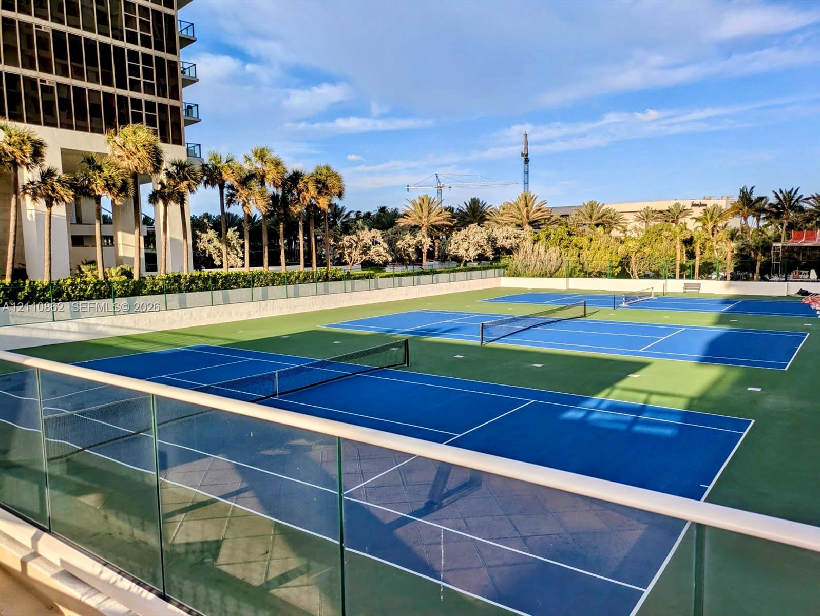 Multiple professional-grade tennis courts featuring blue and green surfaces, perimeter glass panel fencing, and mature palm tree landscaping