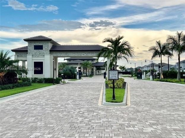 Gated community entrance featuring a stone-clad guardhouse, tile roof, and paver driveway surrounded by mature palm trees and manicured landscaping