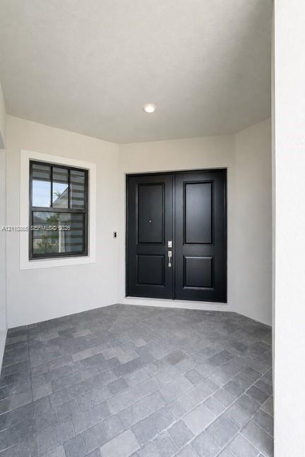 Covered entryway featuring double doors with matte black finish, stone-paver flooring, and a multi-pane window with dark trim Covered entryway featuring double doors with matte black finish, stone-paver flooring, and a multi-pane window with dark trim