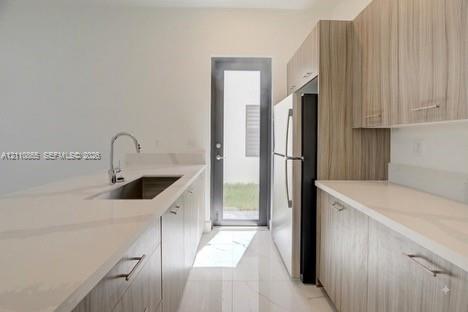 Kitchen featuring flat-panel wood-finish cabinetry, light-toned stone-finish countertops, stainless steel undermount sink with gooseneck faucet, full-height glass-panel exterior door, and polished tile flooring