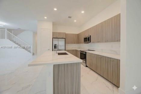 Kitchen featuring flat-panel wood-finish cabinetry, stone-surface countertops, a center island with an integrated sink, stainless steel appliances, and large-format tile flooring Kitchen featuring flat-panel wood-finish cabinetry, stone-surface countertops, a center island with an integrated sink, stainless steel appliances, and large-format tile flooring