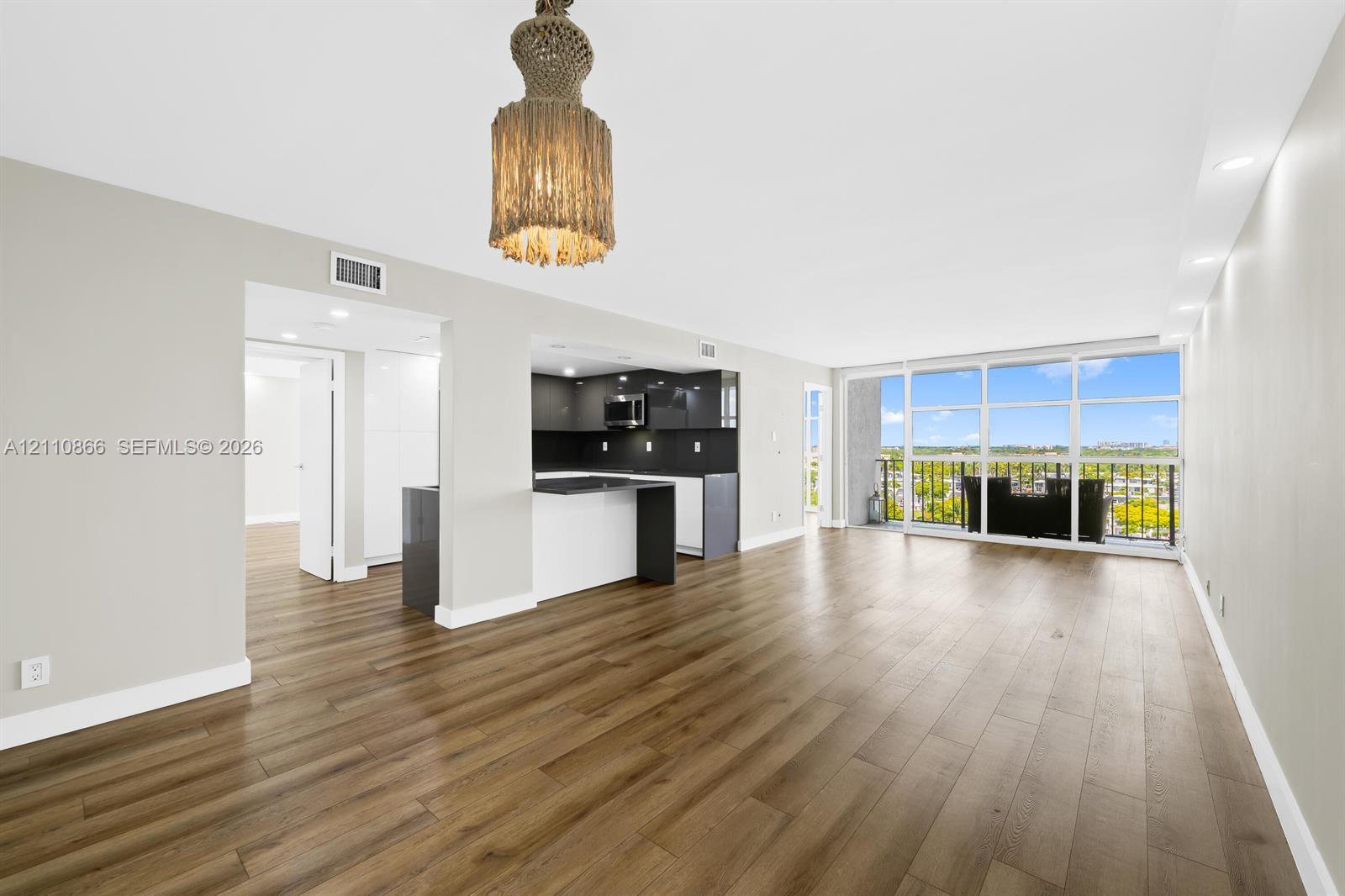 Open-concept space featuring wood-finish flooring, a kitchen with high-contrast cabinetry, recessed lighting, a statement pendant fixture, and floor-to-ceiling windows Open-concept space featuring wood-finish flooring, a kitchen with high-contrast cabinetry, recessed lighting, a statement pendant fixture, and floor-to-ceiling windows