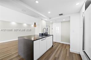 Kitchen featuring white flat-panel cabinetry, a dark-toned island with an integrated sink and gooseneck faucet, wood-finish flooring, and recessed ceiling lighting Kitchen featuring white flat-panel cabinetry, a dark-toned island with an integrated sink and gooseneck faucet, wood-finish flooring, and recessed ceiling lighting