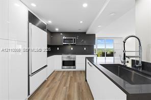 Kitchen featuring high-gloss cabinetry, stainless steel appliances, dark stone-surface countertops, a deep basin sink with a pull-down sprayer faucet, and wood-finish flooring Kitchen featuring high-gloss cabinetry, stainless steel appliances, dark stone-surface countertops, a deep basin sink with a pull-down sprayer faucet, and wood-finish flooring