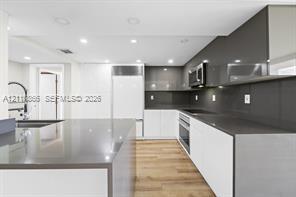 Kitchen featuring white flat-panel cabinetry, dark solid-surface countertops, full-height backsplash, stainless steel built-in appliances, and wood-finish flooring Kitchen featuring white flat-panel cabinetry, dark solid-surface countertops, full-height backsplash, stainless steel built-in appliances, and wood-finish flooring