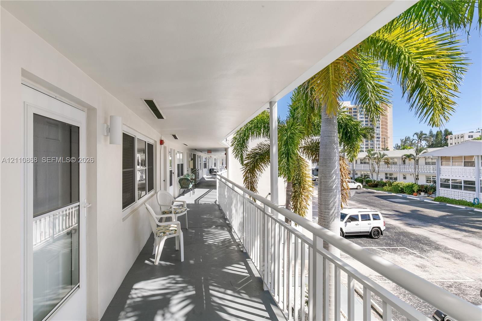 Covered exterior walkway featuring a white metal railing, wall-mounted light fixtures, and solid-surface flooring