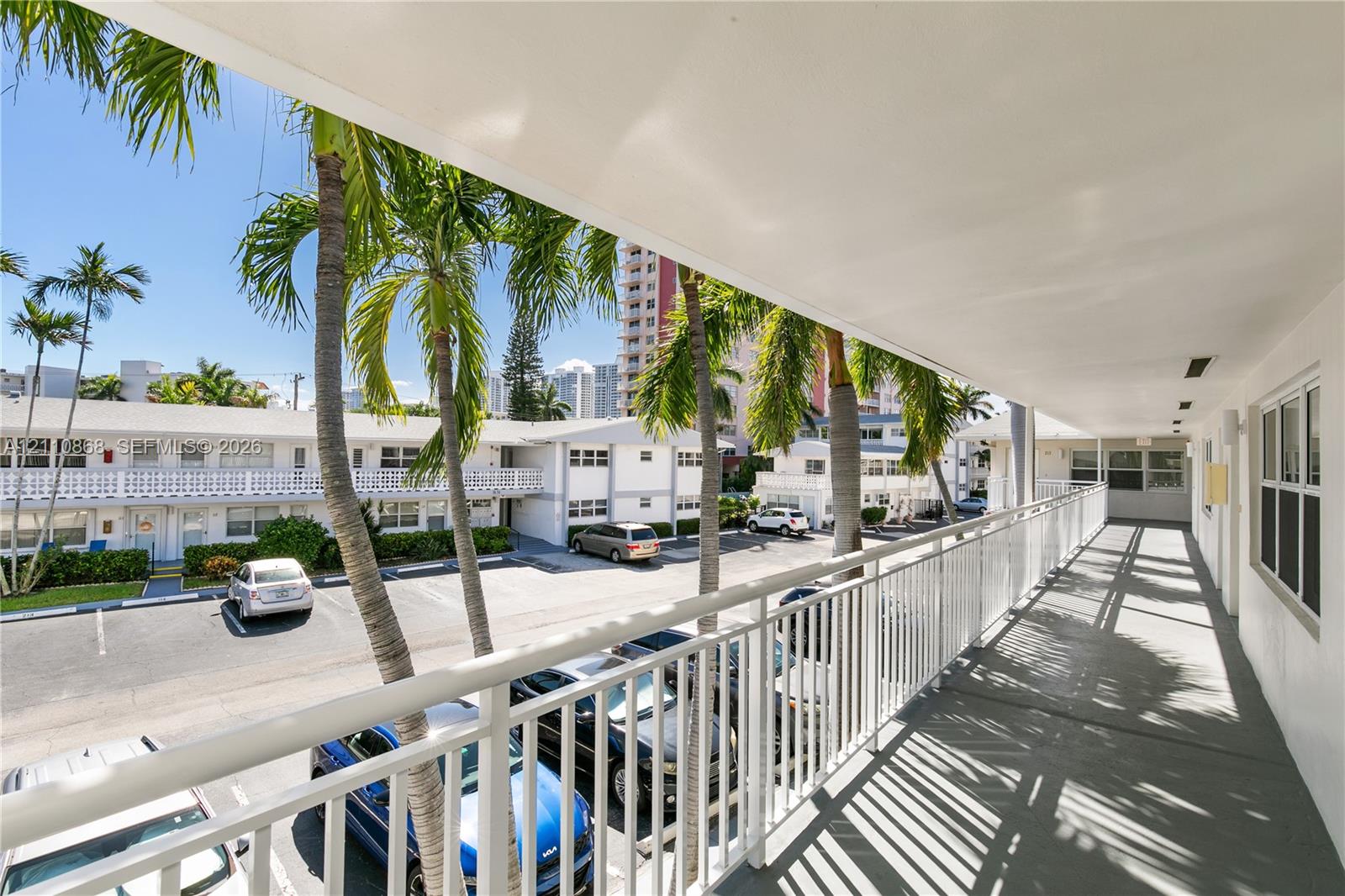Covered exterior walkway featuring white metal railings, concrete flooring, and palm tree landscaping