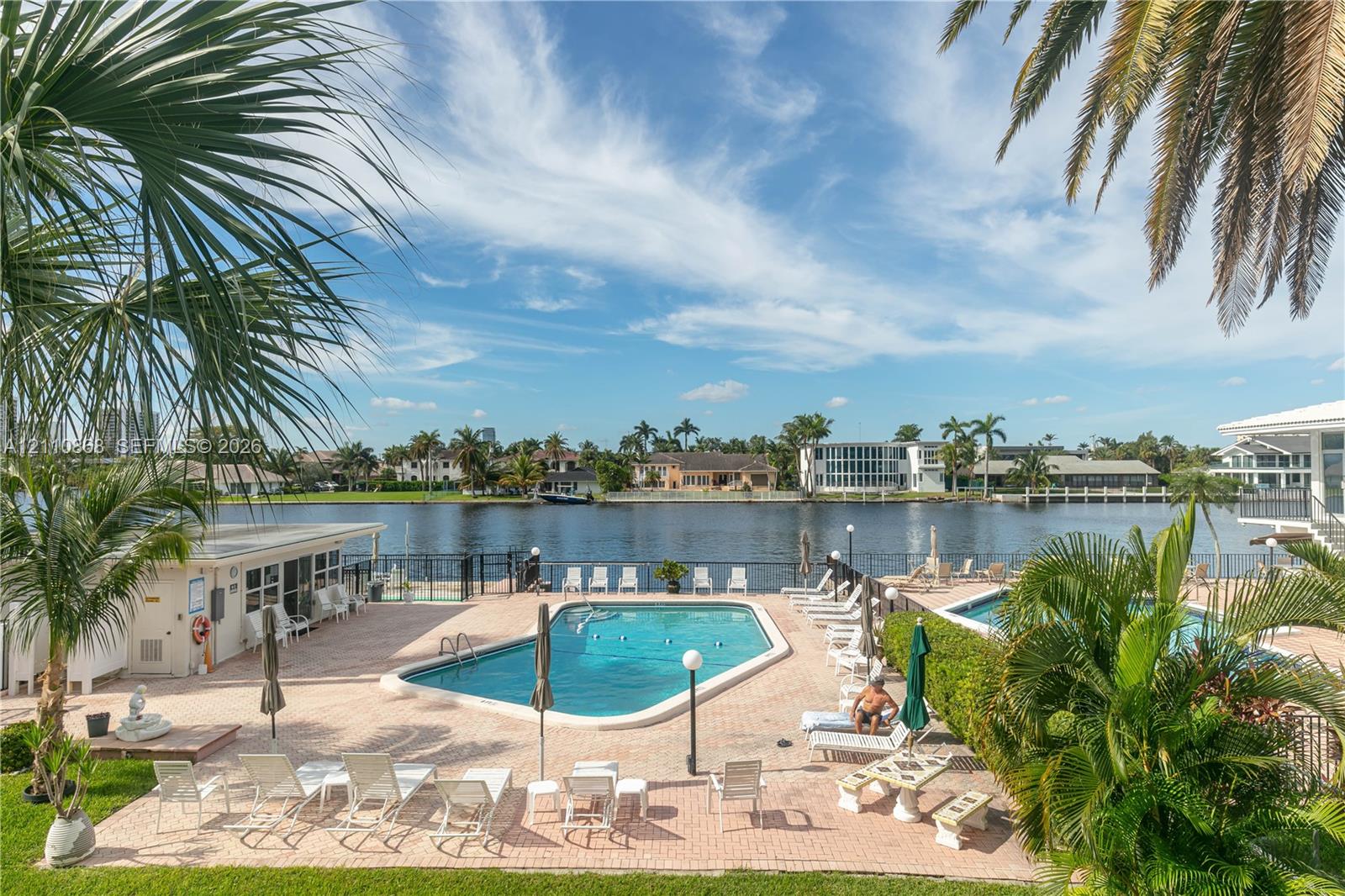 Waterfront pool area featuring a paver deck, metal perimeter fencing, and mature palm trees