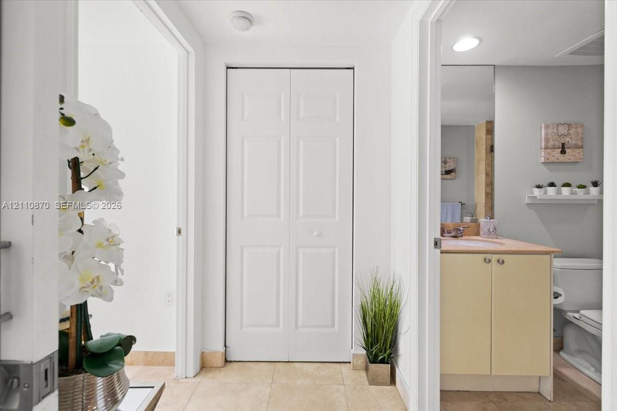 Hallway featuring tile flooring, a white paneled closet door, and an entrance to a bathroom with a vanity, light-toned countertop, and a wall-mounted mirror