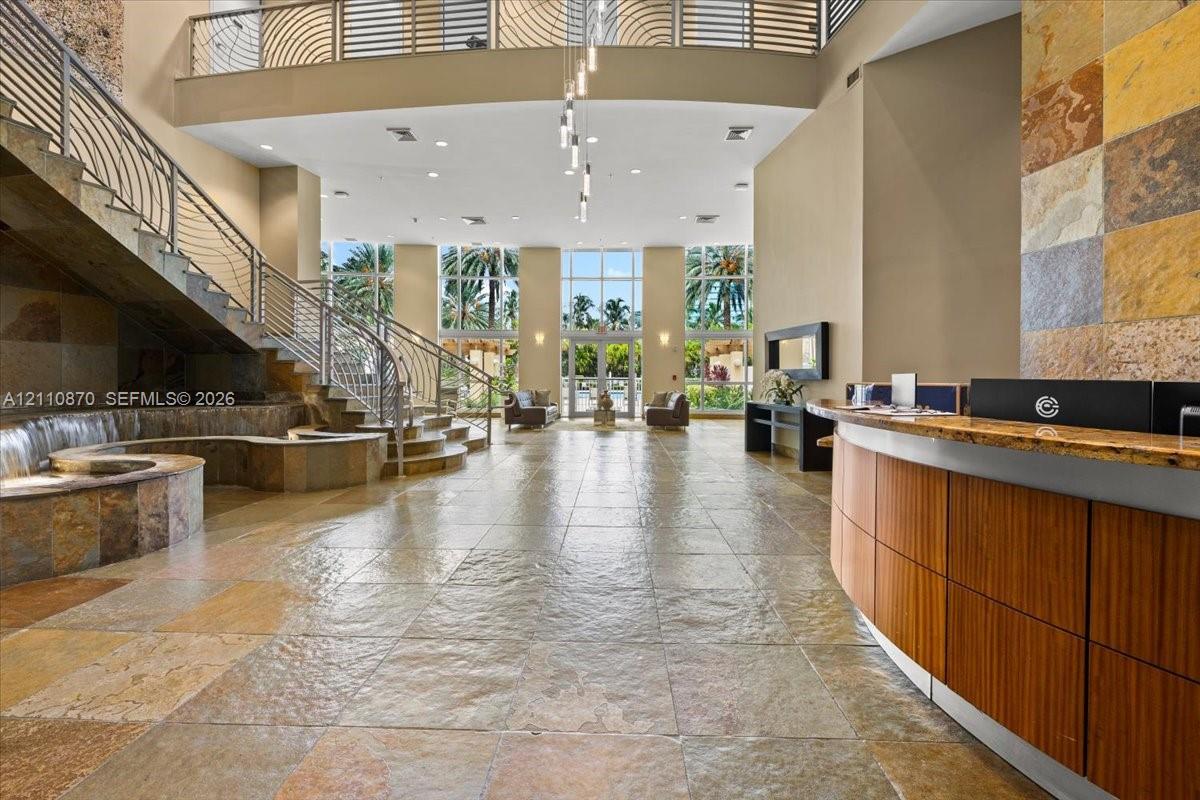 Grand lobby featuring dual-height ceilings, stone tile flooring, a curved wood-panel reception desk, a curved stone staircase with metal railing, and floor-to-ceiling windows