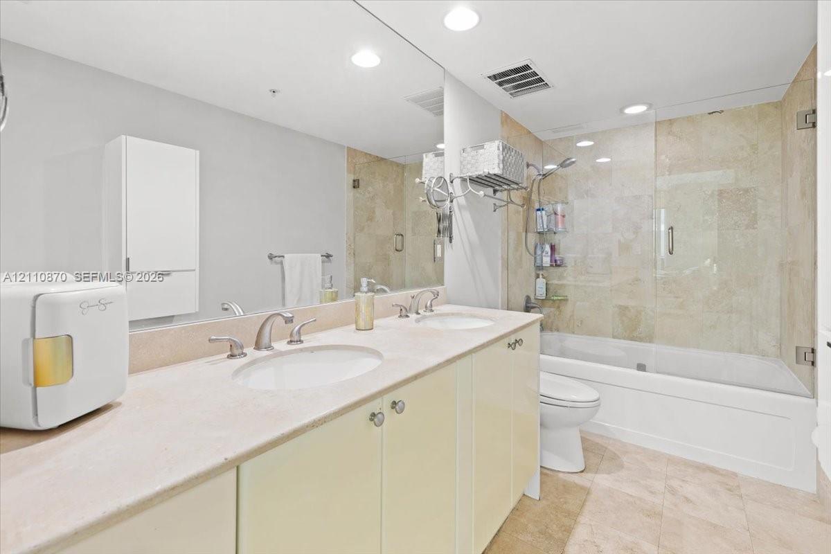 Dual-sink vanity with stone countertop, large wall-to-wall mirror, built-in cabinetry, and a bathtub with glass enclosure and stone-tile surround