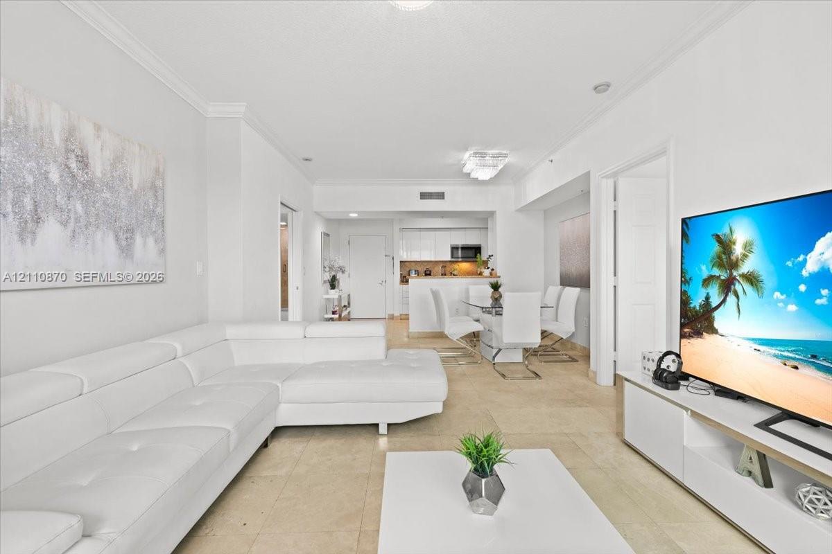 Living area featuring crown molding, neutral tile flooring, and floor-to-ceiling glass sliding doors opening to a private balcony