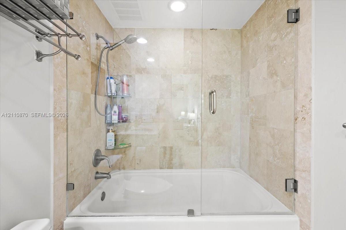 Bathtub with a glass enclosure, wall-mounted shower head, recessed lighting, and tiled wall surround