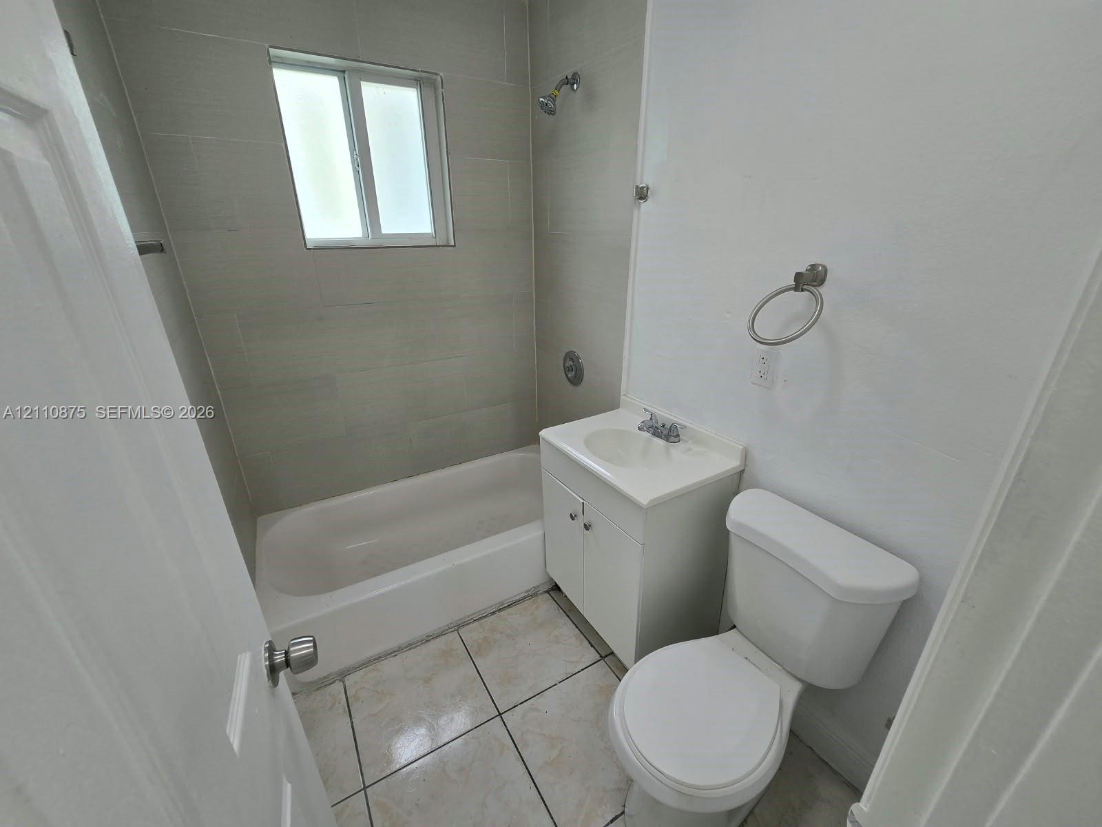 Bathroom featuring light-toned tile flooring, a bathtub with a shower head, wall-mounted shower tile surround, a white vanity cabinet with an integrated basin, and a wall-mounted ring towel holder