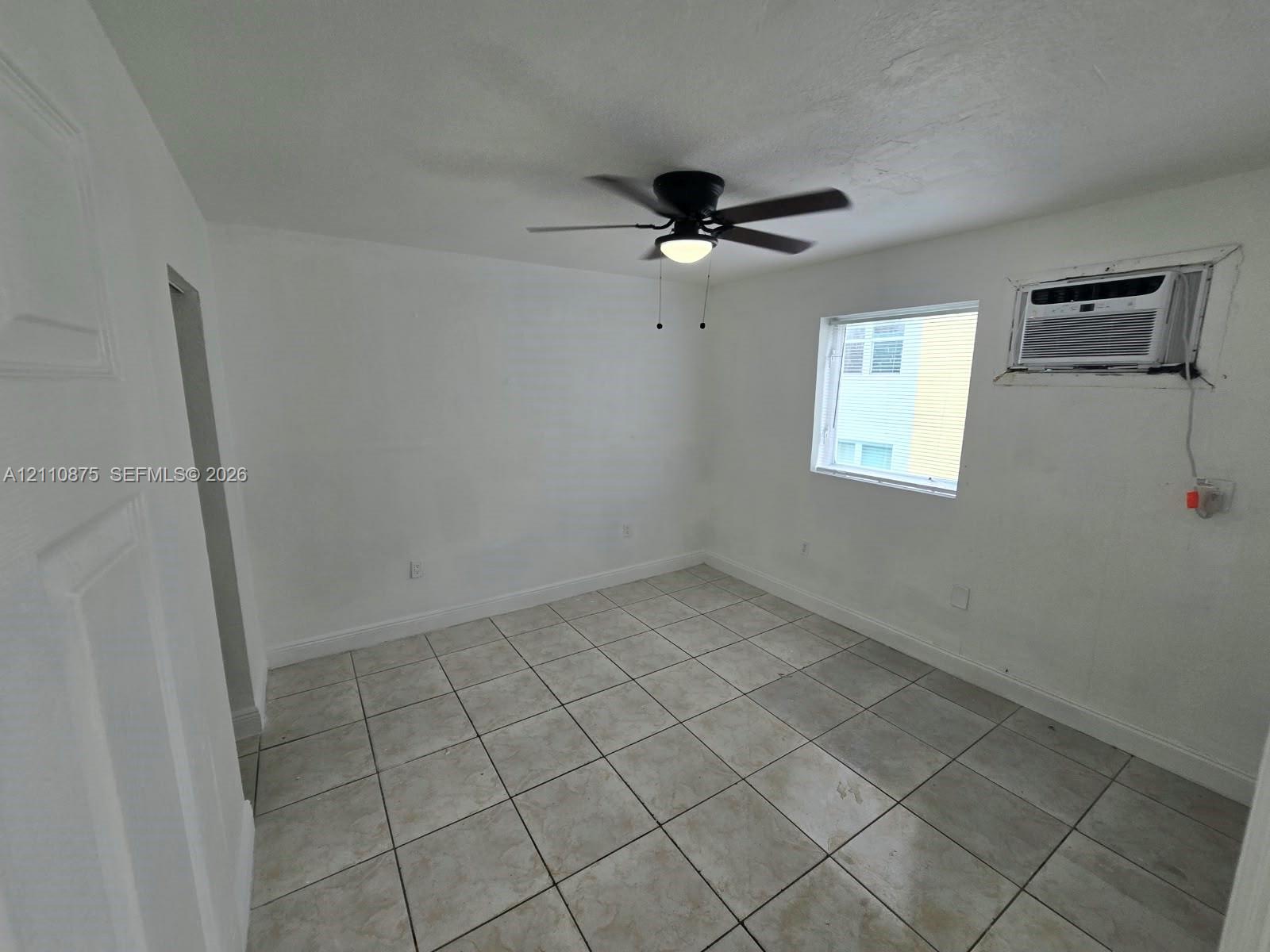 Neutral-toned tile flooring, white wall finishes, black ceiling fan with integrated lighting, and a wall-mounted air conditioning unit