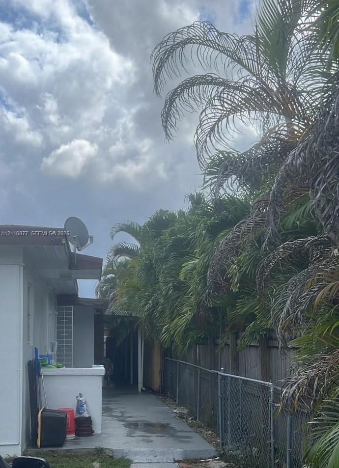 Exterior side walkway featuring painted concrete surfacing, white stucco wall finish, and perimeter chain-link fencing bordered by mature palm trees