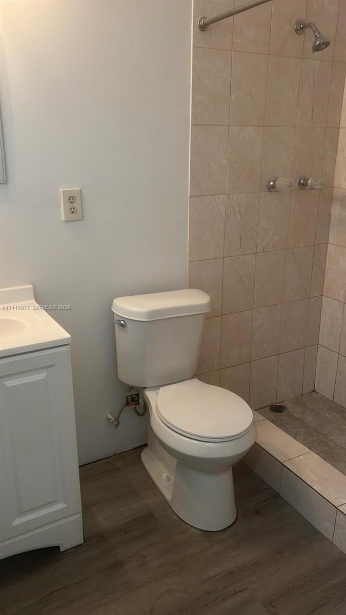 Bathroom featuring wood-finish flooring, a white vanity cabinet, a standard-height toilet, and a shower stall with tiled walls and a metal shower head