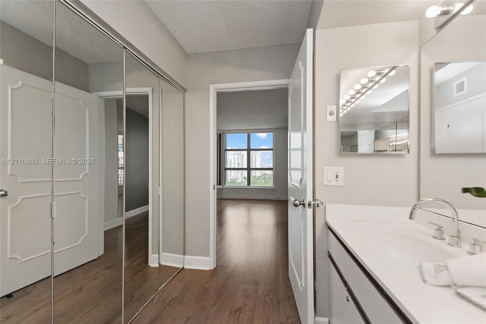 Bathroom featuring a vanity with a white countertop and undermount sink, wall-mounted mirror with integrated lighting, and full-length mirrored closet doors Bathroom featuring a vanity with a white countertop and undermount sink, wall-mounted mirror with integrated lighting, and full-length mirrored closet doors