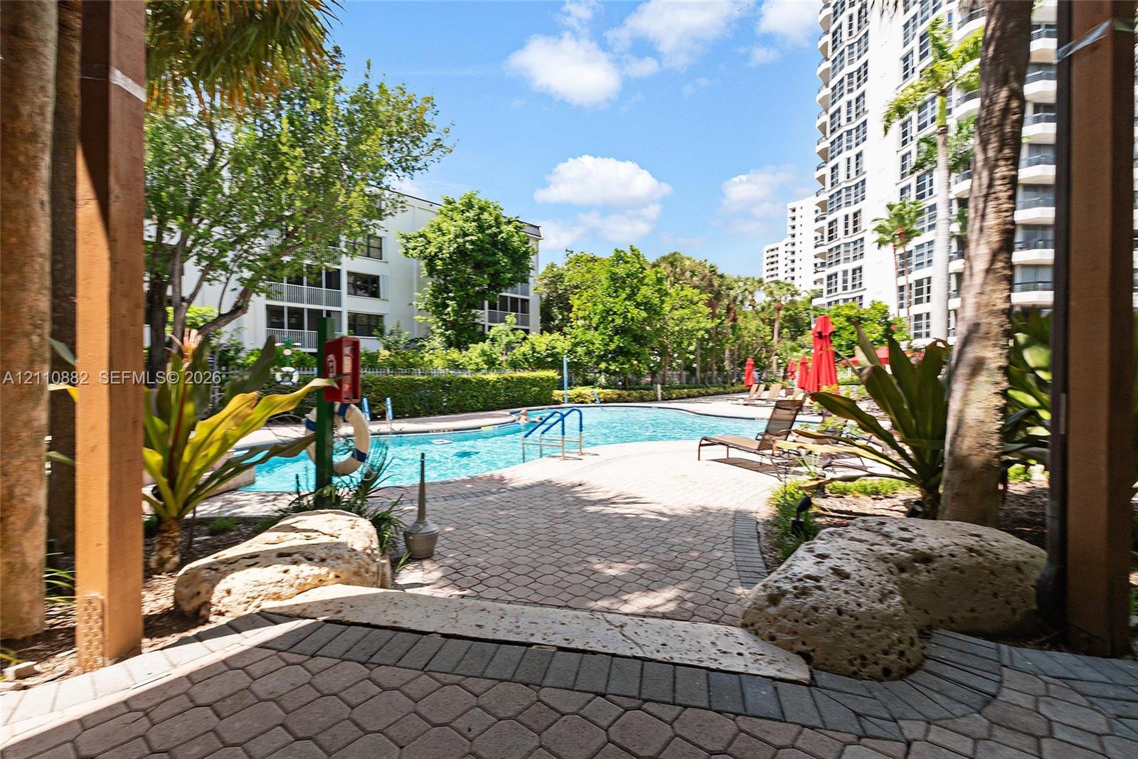 Swimming pool area featuring a paver deck, natural stone boulders, mature palm trees, and lush perimeter landscaping