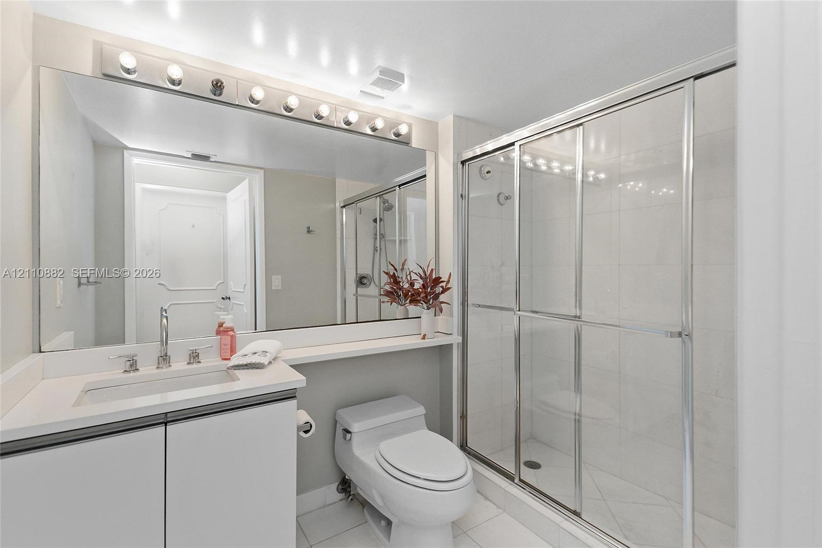Bathroom featuring a vanity with a white countertop and rectangular undermount sink, expansive wall-to-wall mirror, multi-bulb overhead vanity lighting, and a walk-in shower with a glass sliding door and tiled surround