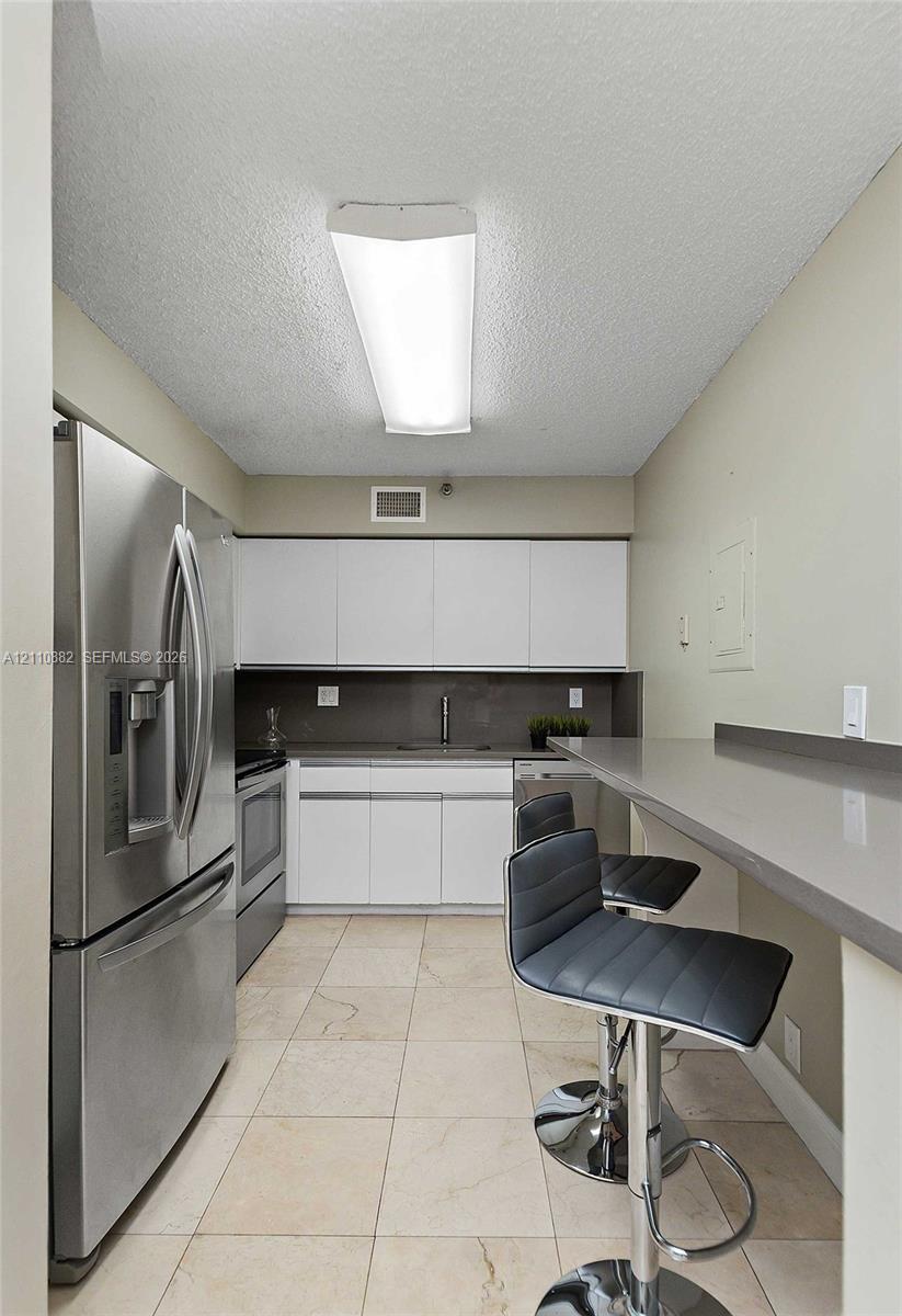 Kitchen featuring white flat-panel cabinetry, stainless steel refrigerator, solid-surface countertop with integrated breakfast bar, and tile flooring Kitchen featuring white flat-panel cabinetry, stainless steel refrigerator, solid-surface countertop with integrated breakfast bar, and tile flooring