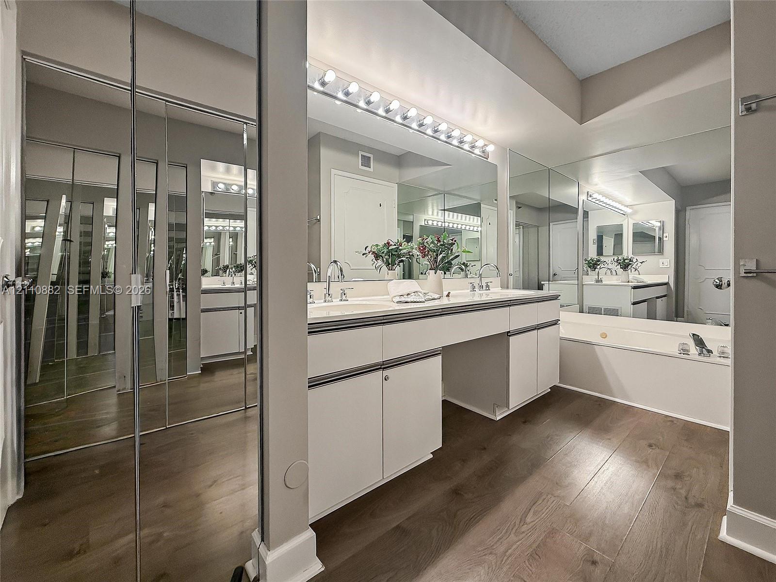 Double vanity featuring white cabinetry, wood-finish flooring, full-wall mirrors, integrated lighting, and a deep soaking tub
