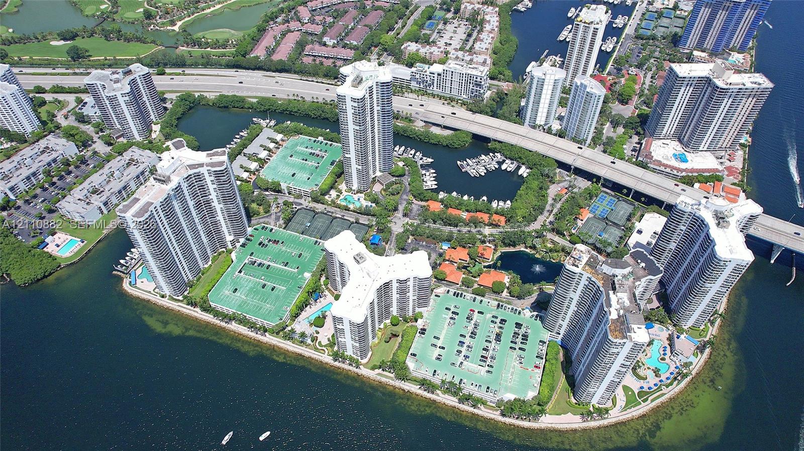 Aerial perspective of waterfront high-rise residential complex featuring multi-level parking structures, multiple swimming pools, and tennis courts situated on a peninsula