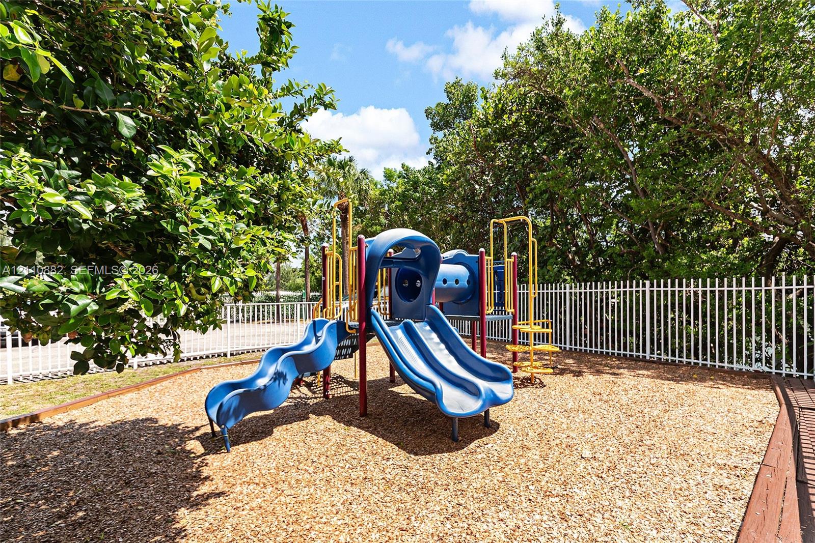 Recreational area featuring a playground structure with dual slides, wood chip ground cover, and a perimeter white metal fence enclosed by mature trees