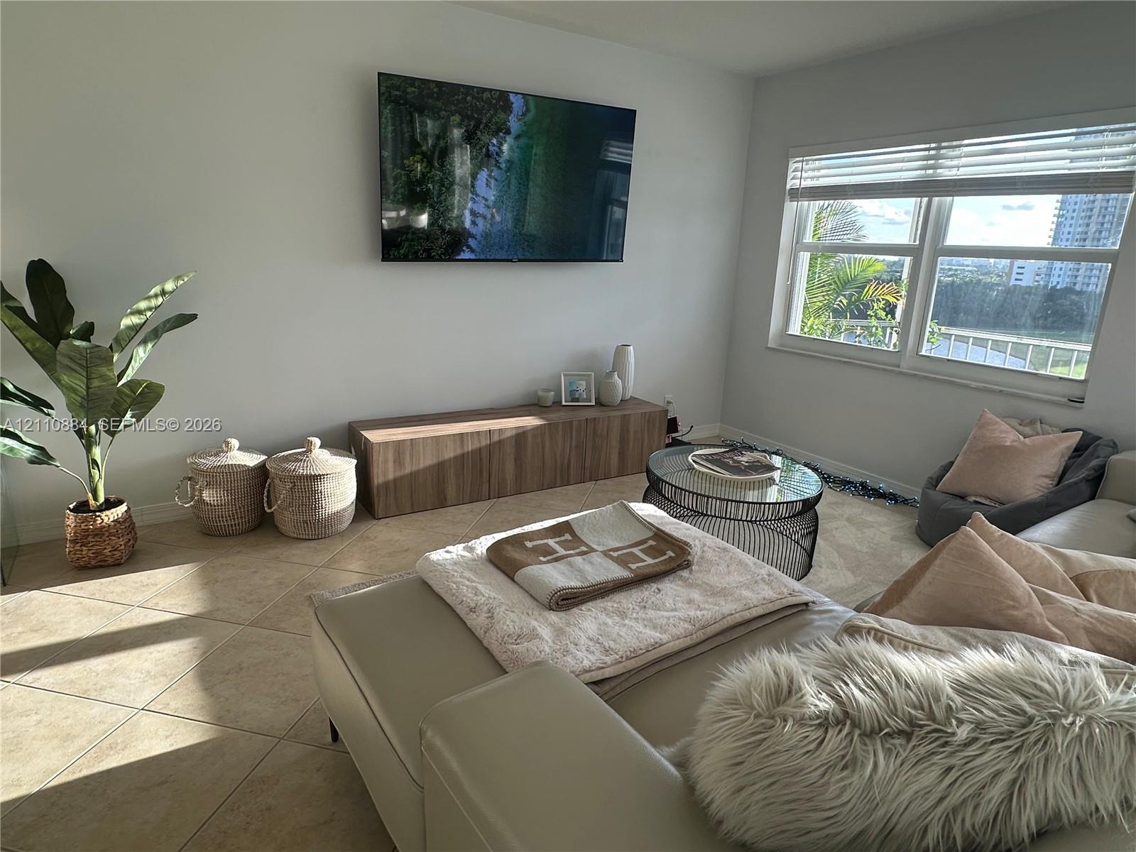 Neutral-tone tile flooring, wall-mounted television, built-in wood-finish cabinetry, and oversized windows with horizontal blinds