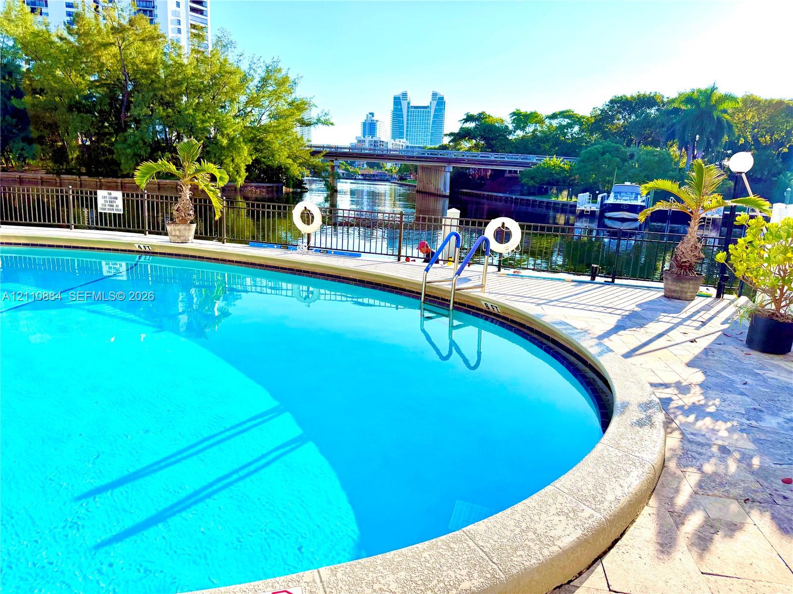 Curved swimming pool featuring stone-paver decking, metal perimeter fencing, and water frontage