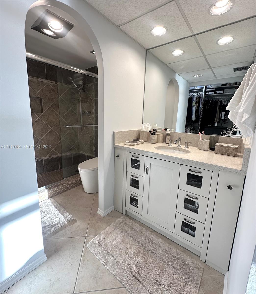 Bathroom featuring a vanity with shaker-style cabinetry, glass-front drawer accents, and a stone-composite countertop with an integrated basin, complemented by an arched doorway, a walk-in shower with tiled surround, and ceramic tile flooring. New ultra-modern style toilet.