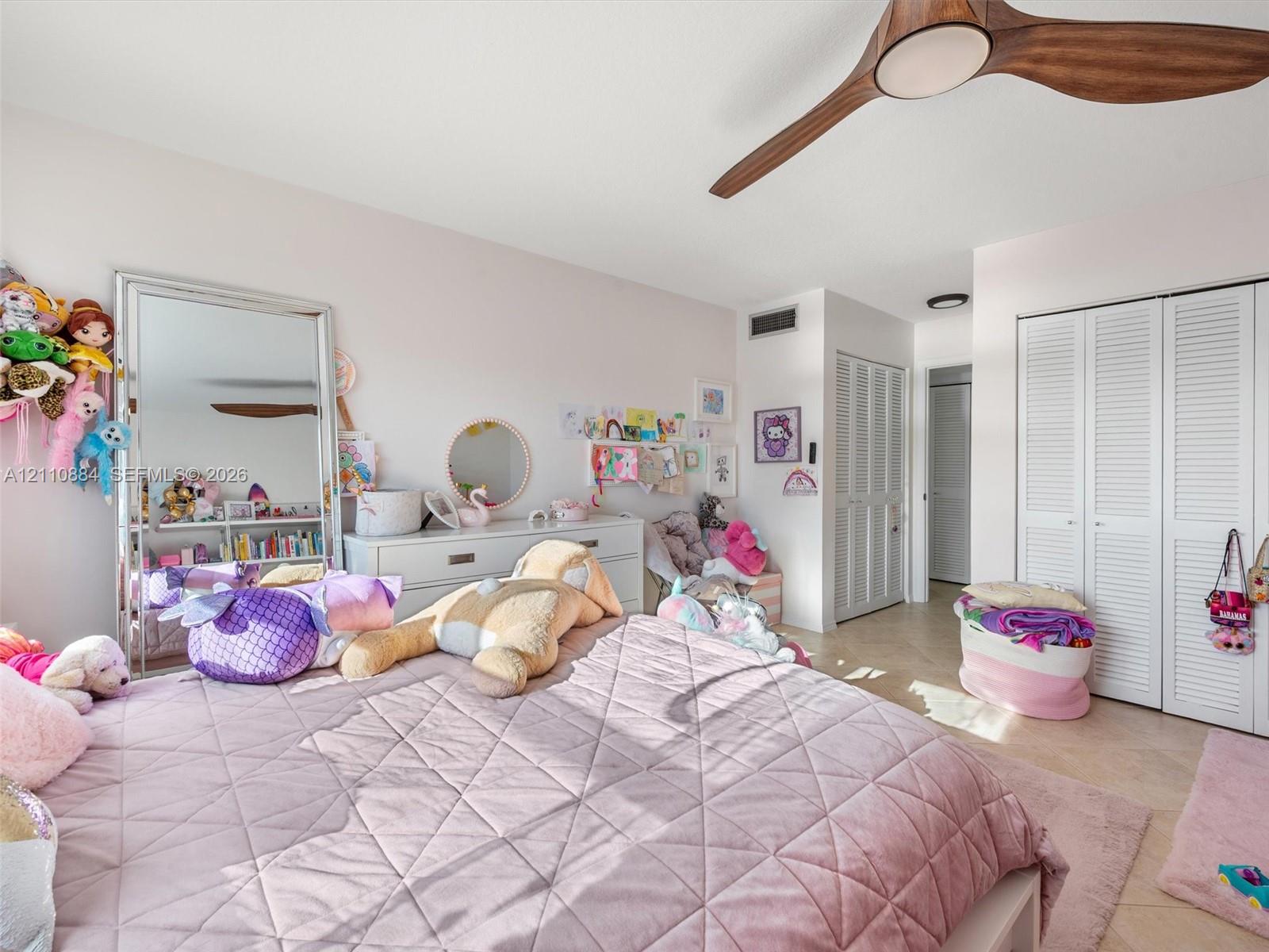 Bedroom featuring neutral-toned tile flooring, dual sets of bi-fold louvered closet doors, and a wood-finish ceiling fan with integrated lighting
