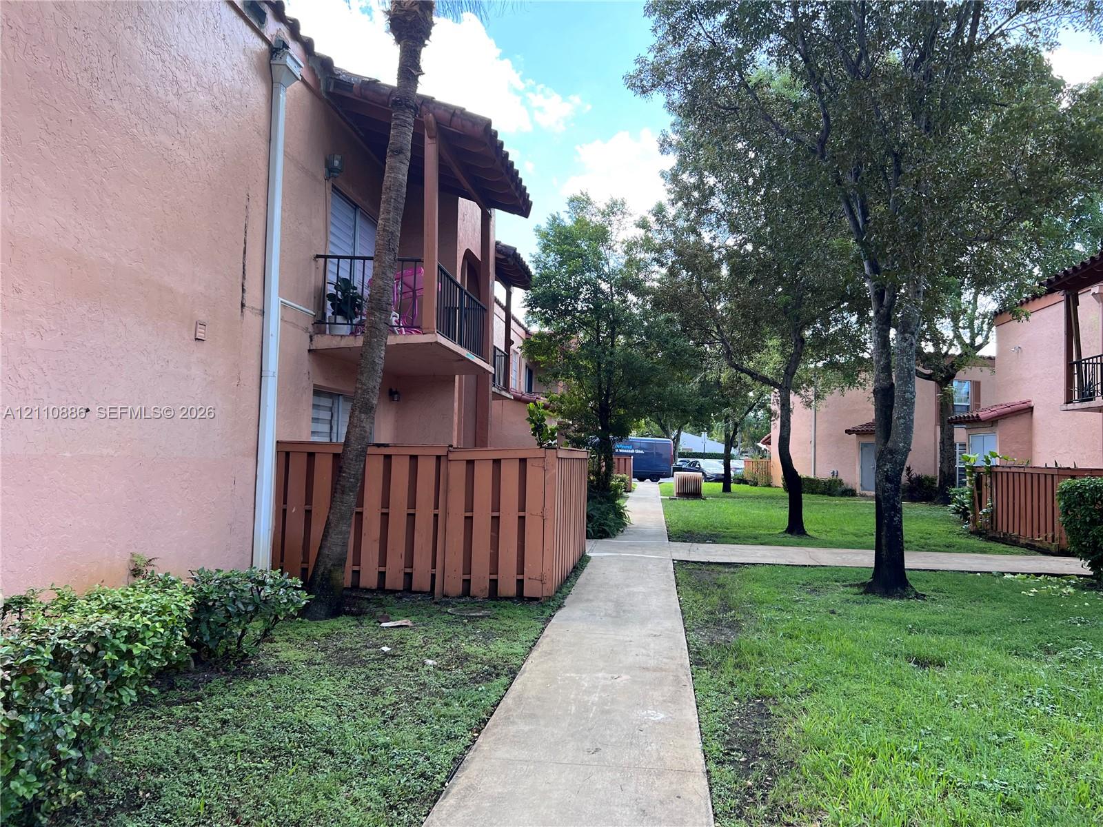 Stucco exterior featuring second-story balconies, concrete walkways, and perimeter wood fencing, situated amidst landscaped grounds with mature trees