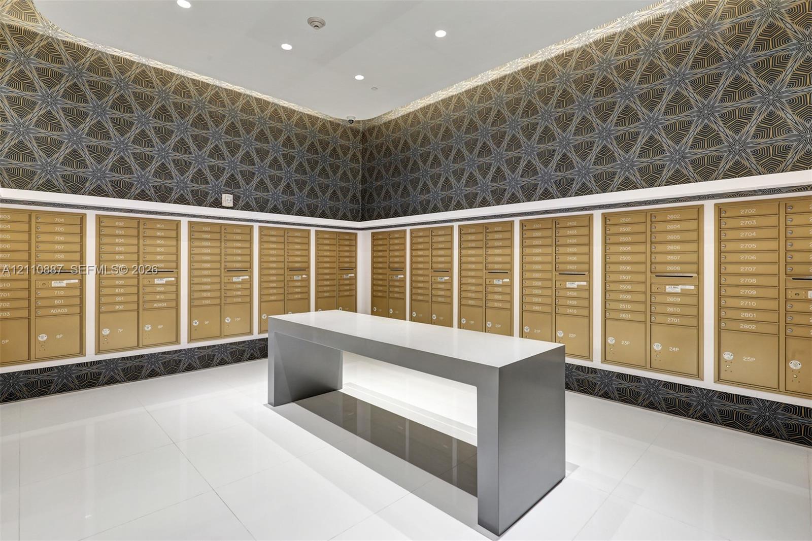 Mail center featuring recessed ceiling lighting, geometric patterned wall covering, polished white floor tiling, and a central freestanding island with a solid surface countertop